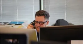 A man who's holding a phone and looking at two monitors, the monitors take up half of the image