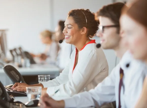 An office with people working all along a row of desks, the image is at a shallow depth of field, which means only the person in the middle of the frame is in focus.