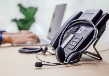 A headset and phone is in the foreground with a laptop and hands in the background.