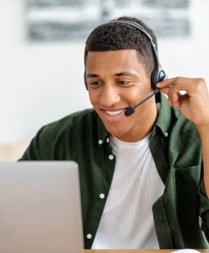 A man smiling while looking at a laptop screen and adjusting his headset.