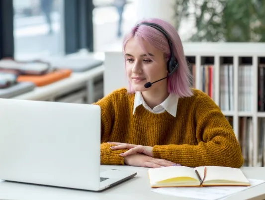 A woman with pink hair sat at a desk looking at a laptop, there is a notebook open next to the laptop.