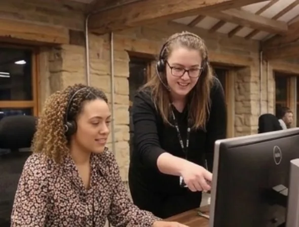 Two women at a desk at the Parallel Partnerships office, one of them is sat down, the other is stood up and pointing at the monitor screen.