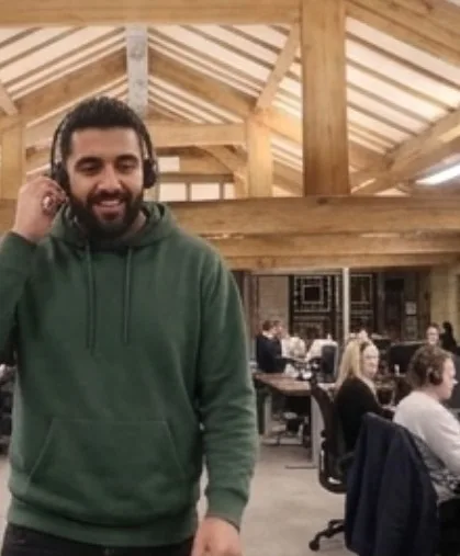 A man stood in the middle of the Parallel Partnerships office with a headset on. Behind him is the rest of the office working at their desks.